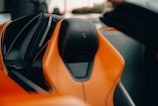 A detailed view of a car seat with a luxurious design, featuring orange leather with black accents and an emblem on the headrest. The background is slightly blurred, indicating the interior of a high-end vehicle.