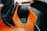 A detailed view of a car seat with a luxurious design, featuring orange leather with black accents and an emblem on the headrest. The background is slightly blurred, indicating the interior of a high-end vehicle.