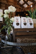 A rustic setting features a bouquet of white roses in a glass vase alongside several paper bags of dog treats on a wooden crate. The background includes wood paneling and hanging lights, enhancing the warm and cozy atmosphere.
