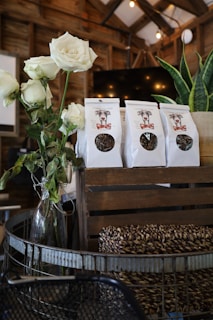 A rustic setting features a bouquet of white roses in a glass vase alongside several paper bags of dog treats on a wooden crate. The background includes wood paneling and hanging lights, enhancing the warm and cozy atmosphere.