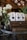 A rustic setting features a bouquet of white roses in a glass vase alongside several paper bags of dog treats on a wooden crate. The background includes wood paneling and hanging lights, enhancing the warm and cozy atmosphere.