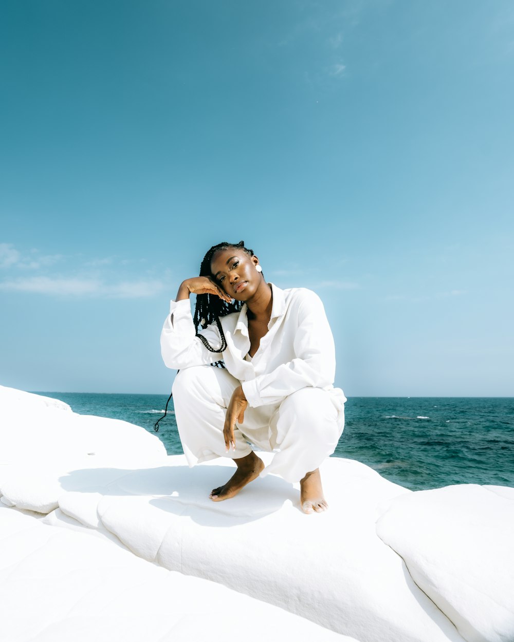 a woman in a white outfit sitting on a rock by the ocean