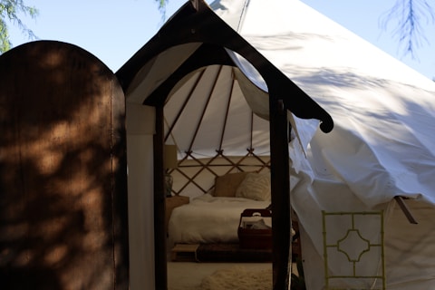A cozy yurt interior is visible through a partially open wooden door. The yurt features a comfortable bed with pillows and a small side table adorned with a decorative plant. Sunlight filters through the canopy, creating soft shadows. The structure showcases a rustic, welcoming atmosphere.