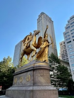 A large golden statue depicting a warrior on horseback accompanied by an angelic figure holding a palm branch. The statue is mounted on a substantial stone pedestal with inscriptions. It is set against the backdrop of tall city buildings and surrounded by trees.