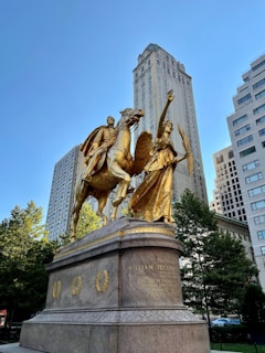 A large golden statue depicting a warrior on horseback accompanied by an angelic figure holding a palm branch. The statue is mounted on a substantial stone pedestal with inscriptions. It is set against the backdrop of tall city buildings and surrounded by trees.