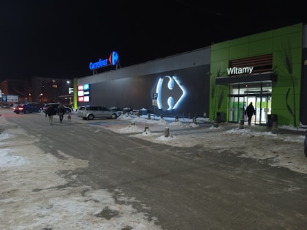 A nighttime scene outside a large Carrefour supermarket with illuminated signage. Several people and cars are visible in the parking lot, and the ground is covered with snow. A green entrance with the word 'Witamy' is seen on the right side.