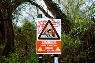 A warning sign is surrounded by dense greenery and trees, indicating that the towpath is flooded and should not be used. The sign features a red and white color scheme with a triangular alert symbol in the center.