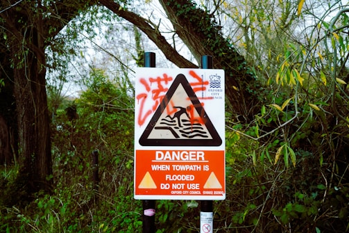 A warning sign is surrounded by dense greenery and trees, indicating that the towpath is flooded and should not be used. The sign features a red and white color scheme with a triangular alert symbol in the center.