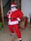 A person dressed in a Santa Claus costume, complete with a red suit, white beard, and Santa hat, is standing indoors. The person is striking a playful pose with sunglasses and hands on hips. There is a large decorative gift box with teddy bear wrapping paper and plants in the background.