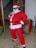 A person dressed in a Santa Claus costume, complete with a red suit, white beard, and Santa hat, is standing indoors. The person is striking a playful pose with sunglasses and hands on hips. There is a large decorative gift box with teddy bear wrapping paper and plants in the background.