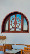 A nautical-themed window features a large ship's wheel incorporated into its design, set against a light gray wall. Below, there is outdoor seating with light blue cushions and wooden frames, along with a small glass-top table.