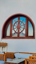 A nautical-themed window features a large ship's wheel incorporated into its design, set against a light gray wall. Below, there is outdoor seating with light blue cushions and wooden frames, along with a small glass-top table.