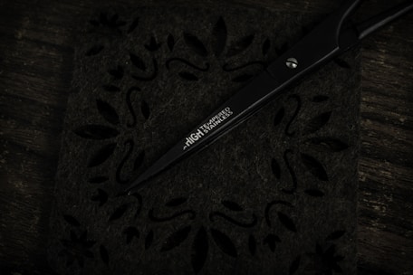 A pair of sleek black scissors rests on a decorative surface with intricate floral and leaf cut-out patterns. The scissors have the words 'HIGH TEMPERED STAINLESS' inscribed on one of the blades. The background and surface are dark, giving a sense of contrast to the metallic sheen of the scissors.