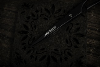 A pair of sleek black scissors rests on a decorative surface with intricate floral and leaf cut-out patterns. The scissors have the words 'HIGH TEMPERED STAINLESS' inscribed on one of the blades. The background and surface are dark, giving a sense of contrast to the metallic sheen of the scissors.