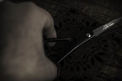A close-up of a hand holding black scissors, cutting a piece of dark fabric with a floral pattern. The lighting is low, creating a moody atmosphere. The scissors have text on the blade indicating a brand.