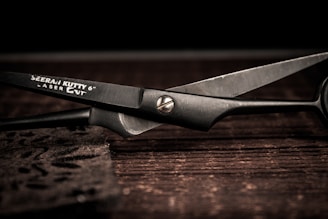 A pair of metal scissors lays open on a dark wooden surface, showcasing sleek black blades and a shiny pivot screw. The brand name and specifications are etched onto one of the blades.