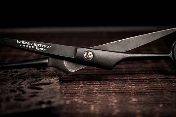 A pair of metal scissors lays open on a dark wooden surface, showcasing sleek black blades and a shiny pivot screw. The brand name and specifications are etched onto one of the blades.