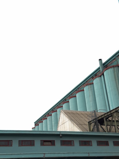 A series of large industrial silos painted in teal stand prominently against the overcast sky. The silos are connected by a network of metal structures and pipes. A building with marked numbers and small windows is visible in the foreground, complementing the industrial aesthetic.