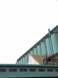 A series of large industrial silos painted in teal stand prominently against the overcast sky. The silos are connected by a network of metal structures and pipes. A building with marked numbers and small windows is visible in the foreground, complementing the industrial aesthetic.