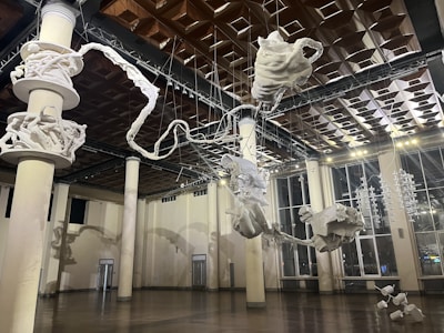 An interior space featuring a large, intricate sculpture suspended from the ceiling. The sculpture appears to be abstract, composed of various white forms resembling twisted ropes and organic shapes. The room is spacious, with tall columns and large windows allowing natural light to filter in. The ceiling is uniquely patterned with geometric designs.
