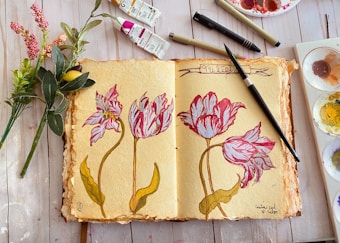 An open sketchbook lies on a wooden table, showing detailed drawings of tulips. The sketches are vibrant with red and white petals and yellow-green stems. Surrounding the sketchbook are drawing tools, including pens and paintbrushes, and tubes of paint. A small bouquet of artificial flowers is placed to the left.