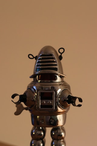 A vintage-style silver robot toy with a dome-shaped head and antenna-like features. The robot has a metallic body with clamp-like hands and jointed legs, standing against a plain background.