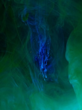 A mysterious and abstract composition of swirling colors with a central focus on deep blues surrounded by shades of green. The textures resemble wisps of smoke or flowing liquid, creating an ethereal and otherworldly atmosphere.