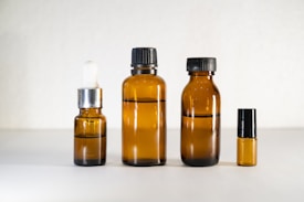 Four amber glass bottles of varying sizes arranged in a row against a neutral background. The bottles include a small dropper bottle, two medium-sized bottles with black caps, and a very small bottle on the far right.