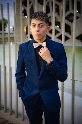 A young person wearing a dark blue suit with a bow tie stands confidently outdoors. The background features a decorative metal railing and a modern architectural structure. The person has a hand slightly adjusting the bow tie and displays a poised demeanor.