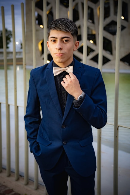 A young person wearing a dark blue suit with a bow tie stands confidently outdoors. The background features a decorative metal railing and a modern architectural structure. The person has a hand slightly adjusting the bow tie and displays a poised demeanor.