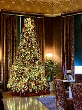 A large, elaborately decorated Christmas tree adorned with golden lights, red ribbons, and gold ornaments stands in a luxurious room. The room features ornate dark curtains, a richly patterned carpet, and elegant furniture, including a finely set dining table with upholstered chairs. A green plant is positioned next to the tree, adding to the festive and opulent atmosphere.
