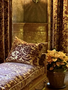 A luxurious interior setting with ornate wall coverings and curtains featuring intricate patterns. A plush, patterned cushion rests on an upholstered couch, also detailed with elaborate designs. A metallic pot holds vibrant orange flowers, adding a touch of color to the scene.