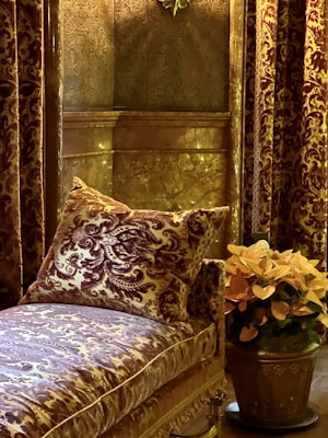 A luxurious interior setting with ornate wall coverings and curtains featuring intricate patterns. A plush, patterned cushion rests on an upholstered couch, also detailed with elaborate designs. A metallic pot holds vibrant orange flowers, adding a touch of color to the scene.