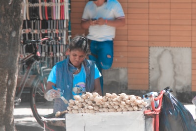 A woman is selling food on a street, sitting next to a cart piled high with what appears to be bags of snacks or produce. Behind her, a person in a white shirt stands against a tiled wall, absorbed in their phone. A bicycle is parked nearby, with numerous belts hanging on display, suggesting a market setting.