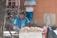 A woman is selling food on a street, sitting next to a cart piled high with what appears to be bags of snacks or produce. Behind her, a person in a white shirt stands against a tiled wall, absorbed in their phone. A bicycle is parked nearby, with numerous belts hanging on display, suggesting a market setting.