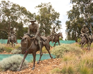 Several bronze statues of soldiers on horseback are positioned in a natural setting with trees and bushes. The soldiers are wearing historical military uniforms and hats, reminiscent of early 20th-century attire. The statues are detailed and life-sized, set against a backdrop of leafy trees and a sky with scattered clouds.