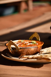 A metal bowl filled with a rich, creamy curry, garnished with white cream swirls and green herbs. The bowl features decorative handles and is placed on a plate alongside pieces of flatbread. The setting conveys a warm, inviting dining atmosphere.