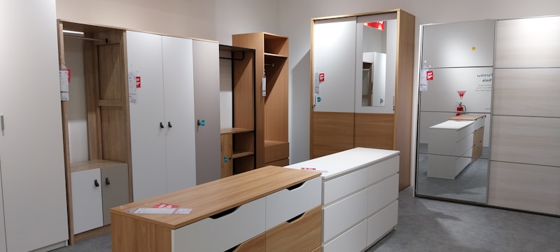 A showroom displaying modern, minimalist wardrobes and dressers with a light wood and white color scheme. The space is organized, showing various styles of wardrobes with sliding and hinged doors, some with mirrors. Tags with product information are visible, suggesting a retail setting.
