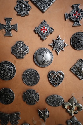 A collection of various metallic emblems and badges with intricate designs, arranged on a tan surface. The emblems feature motifs such as crosses, crests, and Celtic knots, some adorned with red enamel details.