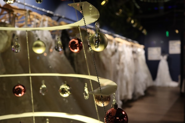 A display showcasing an elegant collection of wedding dresses on hangers. The foreground features a decorative spiral structure adorned with shiny ornaments, including spherical baubles in gold and reddish tones, and crystal-like charms. The background is softly blurred, creating a sense of depth and highlighting the delicate fabrics of the dresses.