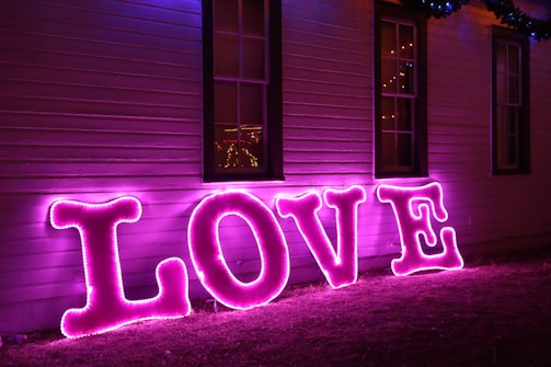 Bright pink neon letters spelling 'LOVE' are placed against a wooden wall, illuminated in a glowing magenta hue. The letters cast a vibrant light that contrasts with the dark exterior of the building.