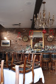 A rustic bar setup with a brick wall backdrop and a large chandelier hanging from the ceiling. The bar counter is lined with various bottles of alcohol, and presents an elegant and vintage look. Chairs are stacked upside down on tables covered with white cloth, indicating the bar is currently closed. Decorative elements like vintage signage and festive ornaments hang from the ceiling, adding charm to the setting.