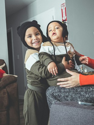 Two young children, dressed in matching outfits with hats featuring animal ears, are smiling and sitting together. They are indoors, possibly in a living room, with a brown couch and a person in the foreground wearing a bright orange sweater.