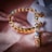 A close-up of a pair of gold hoop earrings intricately embellished with multicolored gemstones in a pattern that resembles eyes. The earrings rest on a soft pink satin surface, with a blurred white background providing contrast.