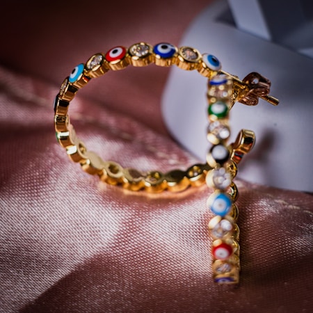 A close-up of a pair of gold hoop earrings intricately embellished with multicolored gemstones in a pattern that resembles eyes. The earrings rest on a soft pink satin surface, with a blurred white background providing contrast.