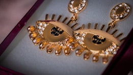 A pair of gold earrings designed in the shape of stylized eyes, adorned with sparkling crystals and decorative elements resembling eyelashes. The earrings are set in a jewelry box with a soft, velvet-like interior.