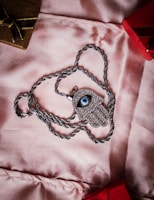 A silver necklace with a pendant shaped like an eye, encrusted with small stones, lies on a soft pink fabric. The chain is silver and twisted, forming a loop around the pendant. In the background, there are parts of two small boxes, one brown with a golden ribbon and the other red.