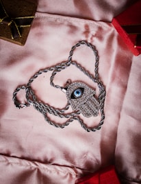 A silver necklace with a pendant shaped like an eye, encrusted with small stones, lies on a soft pink fabric. The chain is silver and twisted, forming a loop around the pendant. In the background, there are parts of two small boxes, one brown with a golden ribbon and the other red.