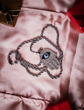 A silver necklace with a pendant shaped like an eye, encrusted with small stones, lies on a soft pink fabric. The chain is silver and twisted, forming a loop around the pendant. In the background, there are parts of two small boxes, one brown with a golden ribbon and the other red.
