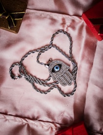 A silver necklace with a pendant shaped like an eye, encrusted with small stones, lies on a soft pink fabric. The chain is silver and twisted, forming a loop around the pendant. In the background, there are parts of two small boxes, one brown with a golden ribbon and the other red.